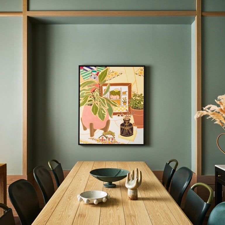 A long wooden dining table with decorative bowls and a hand sculpture sits under a colorful framed artwork on a sage green paneled wall. "HOW TO LIVE WITH OBJECTS" and "THE ANIMAL KINGDOM" books are visible on a side console.