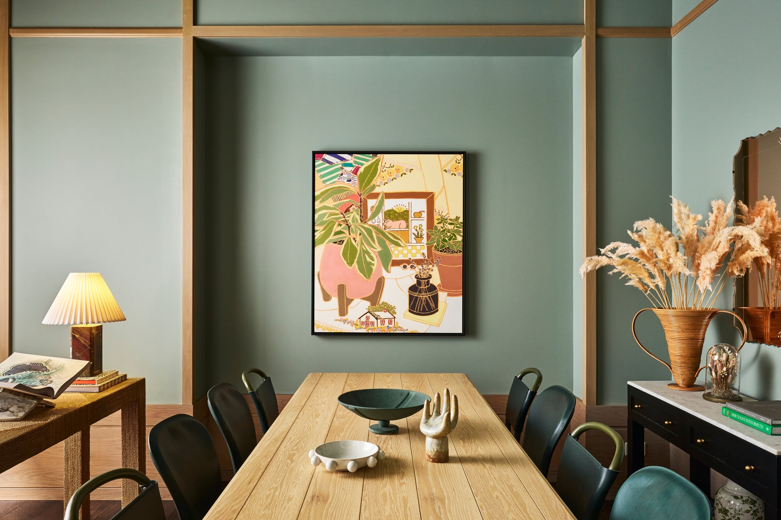 A long wooden dining table with decorative bowls and a hand sculpture sits under a colorful framed artwork on a sage green paneled wall. "HOW TO LIVE WITH OBJECTS" and "THE ANIMAL KINGDOM" books are visible on a side console.
