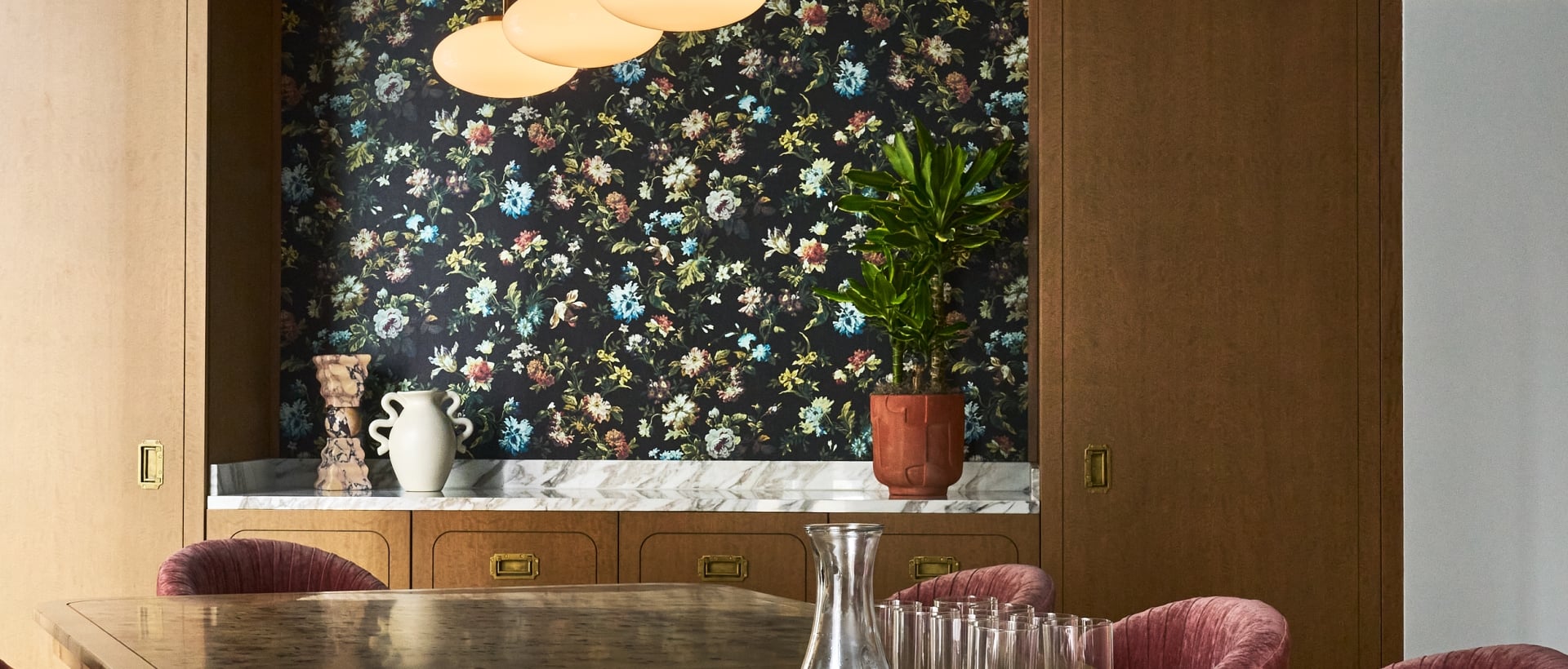 A burlwood dining table displays a wicker tray, carafe, and glasses, surrounded by red chairs. Brass pendant lights hang from a teal ceiling over a floral wallpaper backdrop.