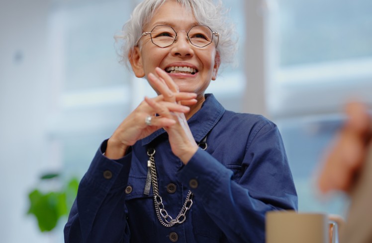 An elderly Asian woman with grey hair and glasses smiles broadly, hands clasped, wearing a dark blue shirt with a silver chain, seated at a table indoors.