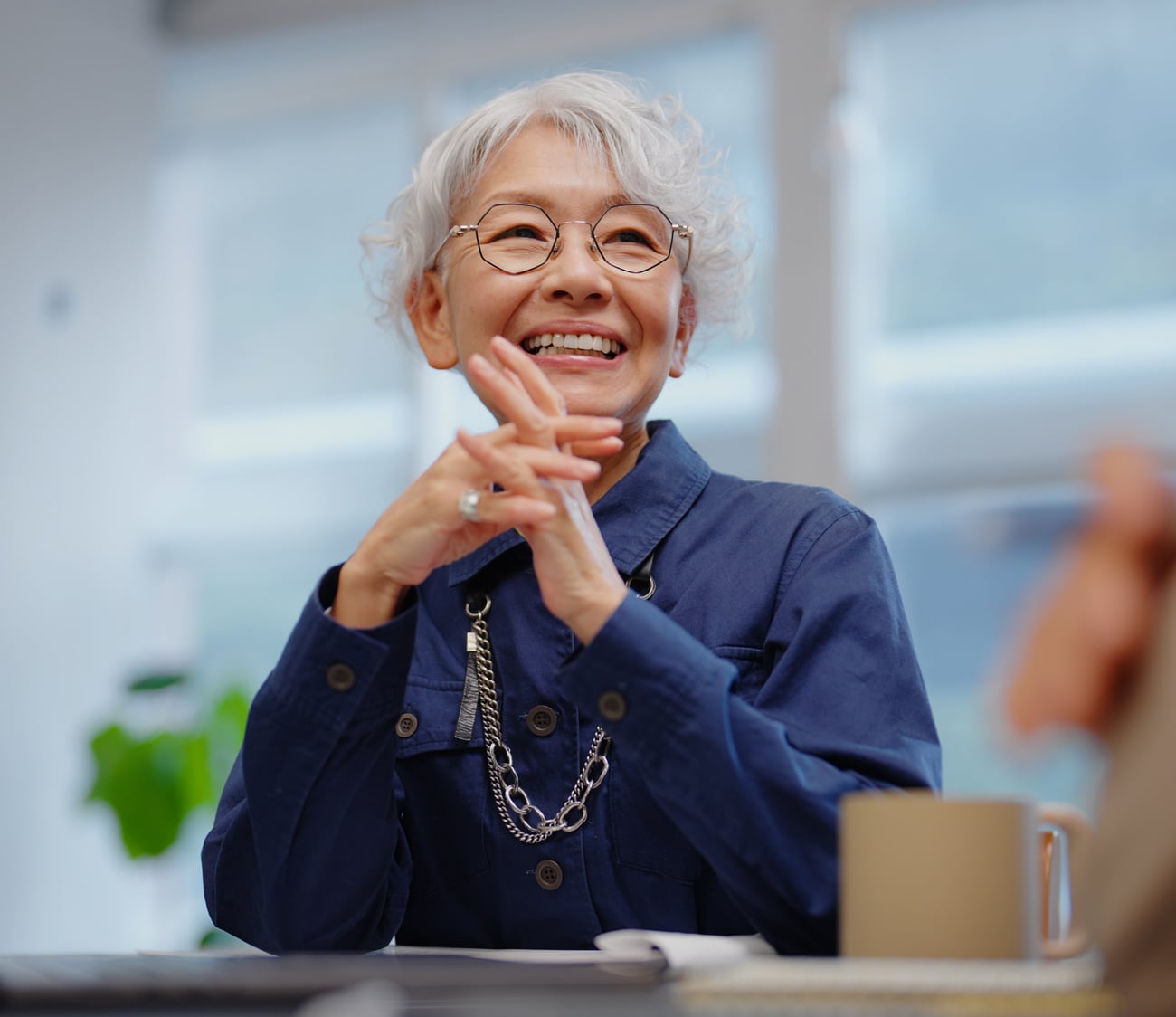 An elderly Asian woman with grey hair and glasses smiles broadly, hands clasped, wearing a dark blue shirt with a silver chain, seated at a table indoors.