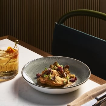 A bowl of fried food items with mushrooms and diced red pieces in sauce, and a cocktail, sit on a marble table with cutlery, against a dark paneled wall.