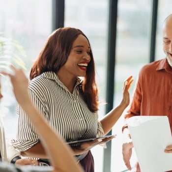 A Black woman in a striped shirt laughs, holding a tablet and gesturing, with a smiling bald man holding papers and other colleagues nearby in a bright office.