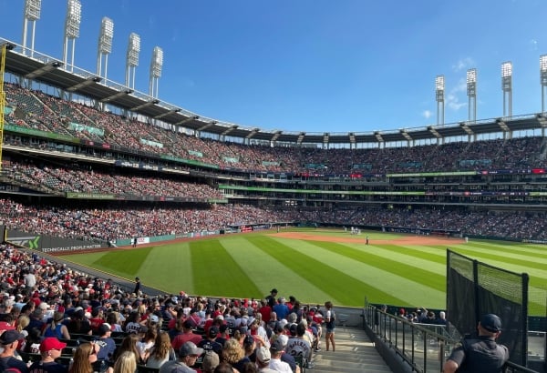 A crowded baseball stadium hosts a game on a sunny day, viewed from spectator seats overlooking the field.
