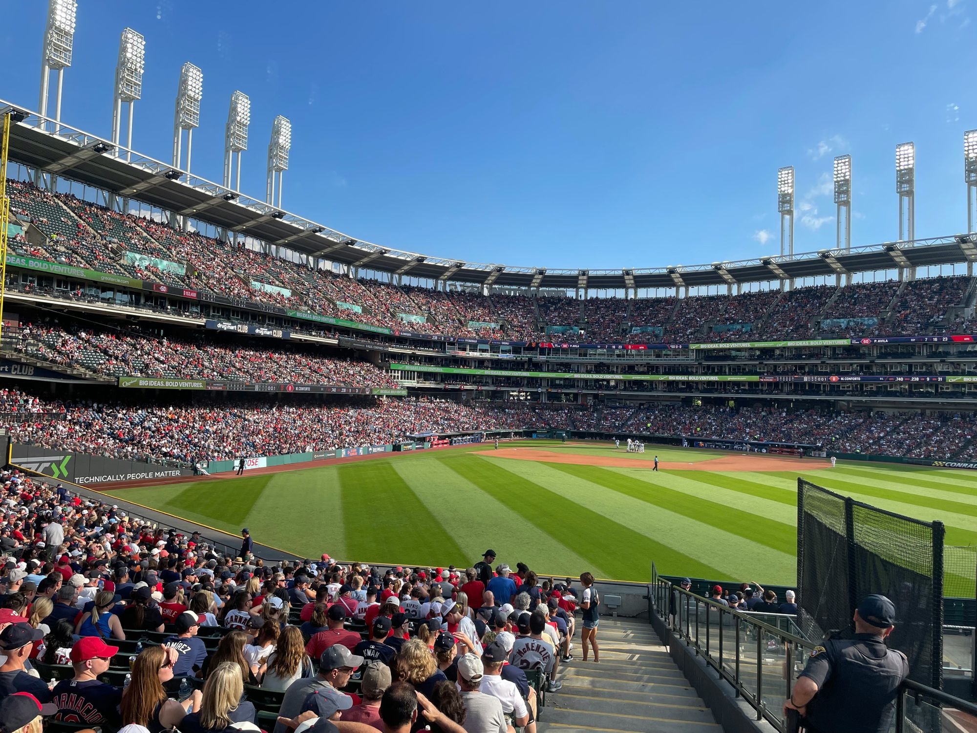 A crowded baseball stadium hosts a game on a sunny day, viewed from spectator seats overlooking the field.