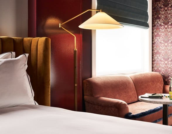 A brass floor lamp illuminates a hotel room with a gold velvet headboard, a pink velvet loveseat, and patterned wallpaper next to a brightly lit window.