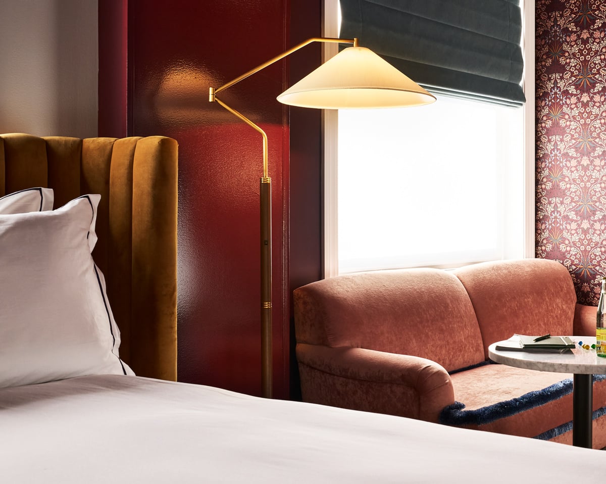 A brass floor lamp illuminates a hotel room with a gold velvet headboard, a pink velvet loveseat, and patterned wallpaper next to a brightly lit window.