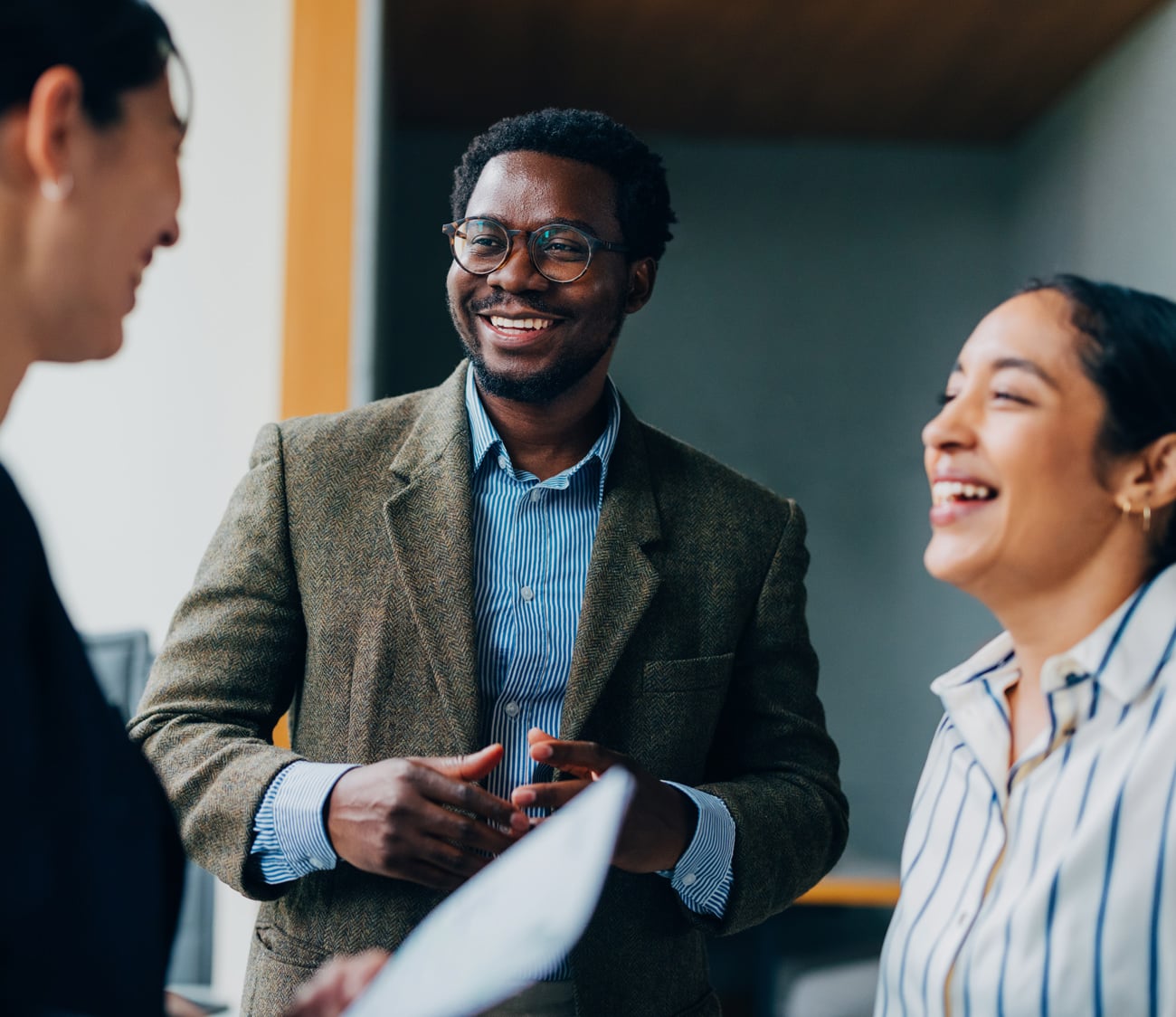 A smiling Black man in glasses talks, gesturing with his hands, with two laughing women on either side in an office setting.