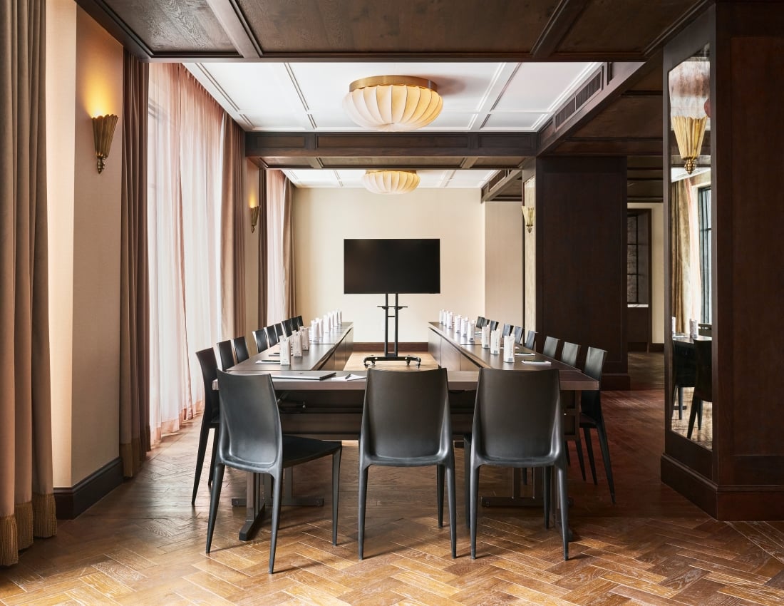 Long conference table with black chairs, set for a meeting, features a large screen and cartons labeled "JUST STILL WATER 500ml". Room has a herringbone floor, dark wood accents, and light curtains.