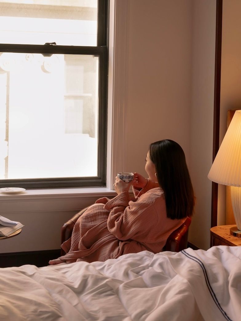 A woman sits in an armchair, holding a patterned teacup and looking out a large window. She is in a softly lit bedroom with a bed and a pleated bedside lamp.
