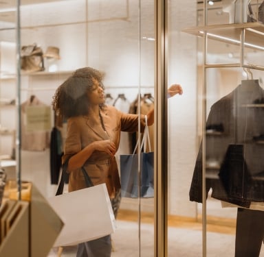 A woman with curly hair, holding shopping bags, looks into a luxury clothing store's glass display. She is reaching towards items on a rack inside the store.