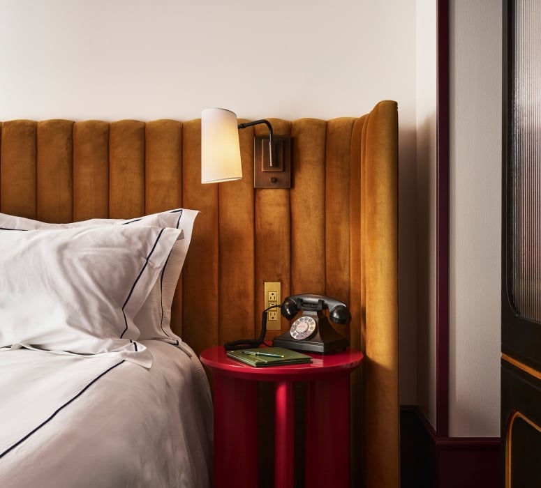 A bed with a mustard velvet headboard and white bedding; a black vintage rotary phone and green notebook rest on a red cylindrical nightstand. A wall sconce hangs above.