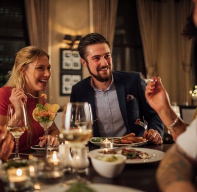 A laughing woman and smiling man eat and drink wine at a dimly lit restaurant table set with candles. Blurred city lights are outside.