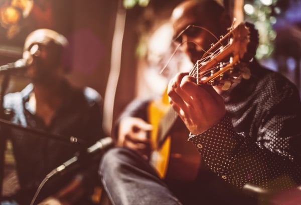 A musician's hands play an acoustic guitar's fretboard, wearing a polka-dot shirt. Another blurred musician stands nearby amidst warm, bokeh lights.