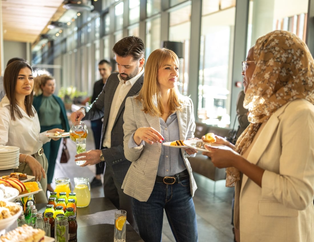 People are holding plates and drinks, engaging in conversation around a buffet table laden with food and beverages at a bright, modern indoor gathering.