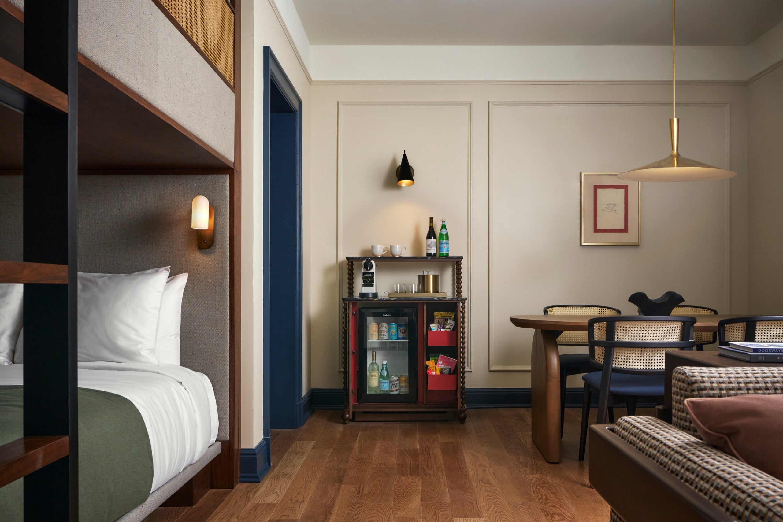 A hotel room contains a bunk bed, a minibar (with 'Innovative' fridge, 'CIGAR CITY', '50', 'S.PELLEGRINO', 'Cincinnati Light' drinks), and a dining table with rattan chairs. Books 'DOORWAYS', 'CIRCLE' are visible.
