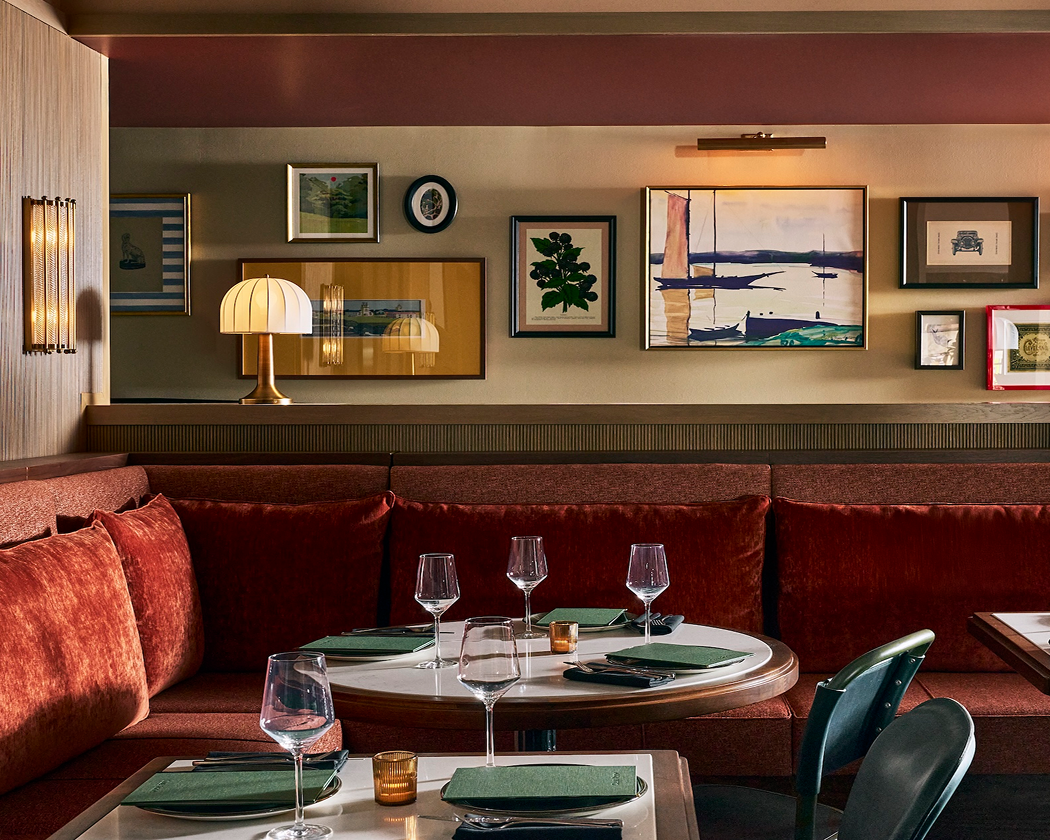 Red velvet banquette seating and white marble tables set with glasses in a restaurant. A beige wall displays varied framed artworks and a gold table lamp.