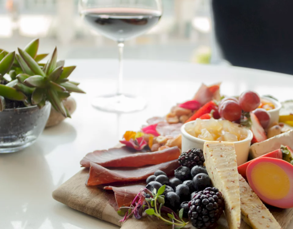 A charcuterie board presents cured meats, cheeses, berries, fruits, and a pink-dyed egg, set on a white table with a glass of red wine and a succulent plant.