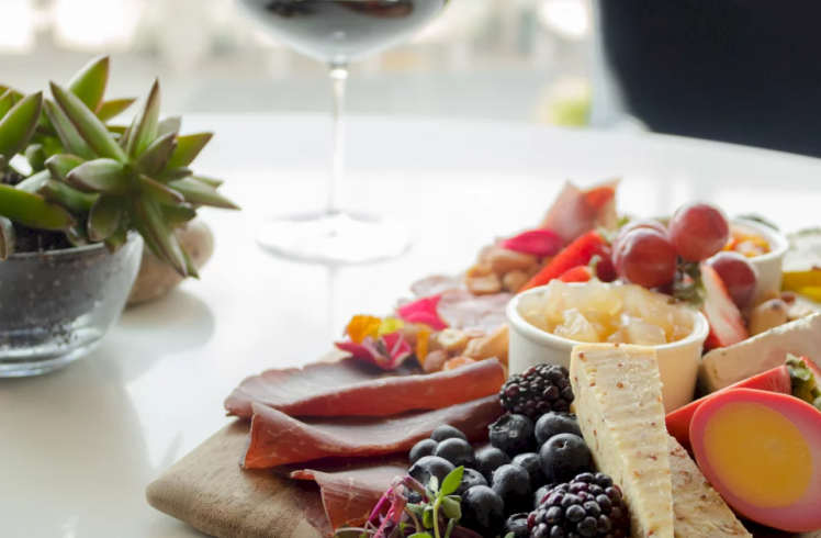 A charcuterie board presents cured meats, cheeses, berries, fruits, and a pink-dyed egg, set on a white table with a glass of red wine and a succulent plant.