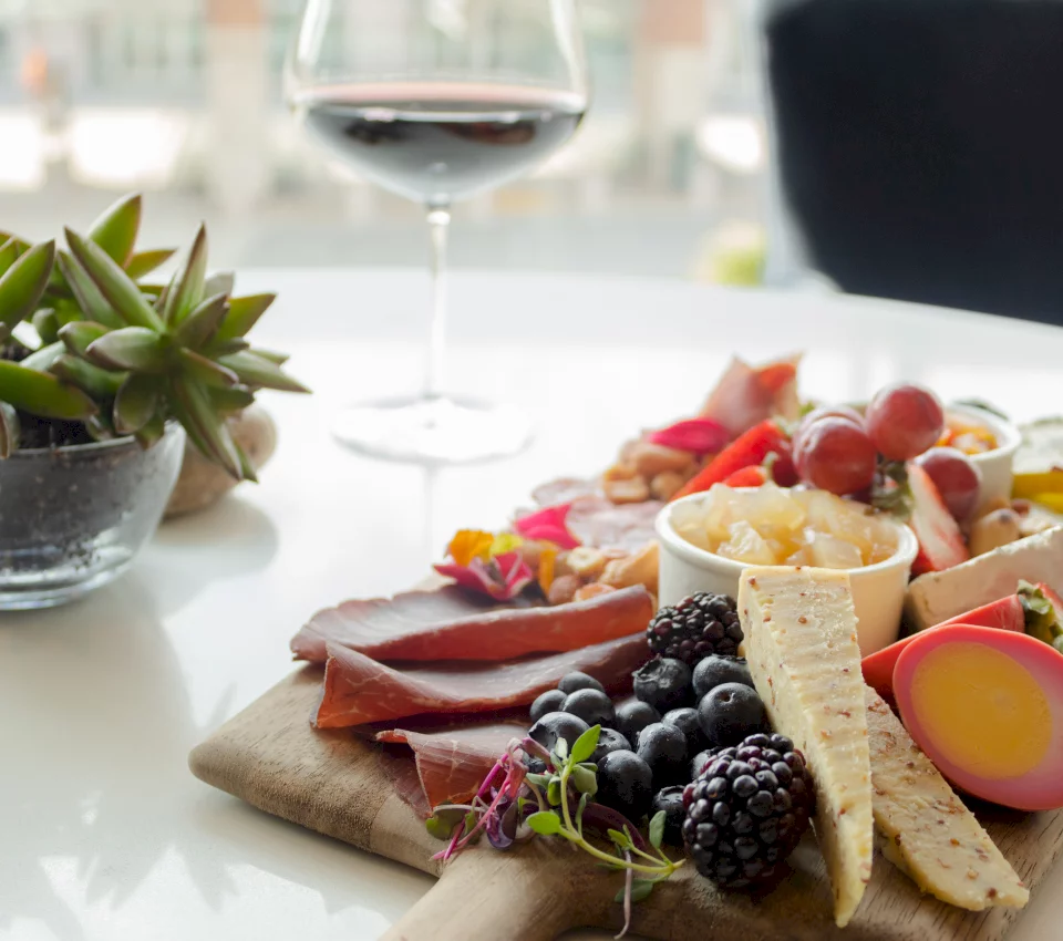 A charcuterie board presents cured meats, cheeses, berries, fruits, and a pink-dyed egg, set on a white table with a glass of red wine and a succulent plant.