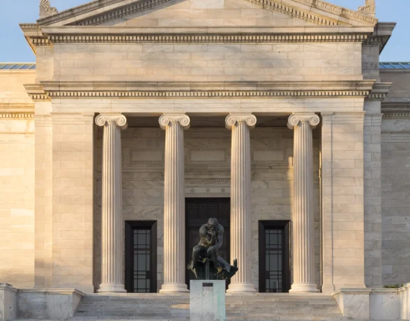 A bronze statue of a pensive, nude man sits on a pedestal at the base of a wide staircase leading to a grand classical stone building with four columns.