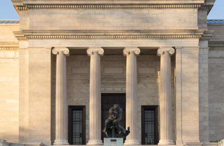 A bronze statue of a pensive, nude man sits on a pedestal at the base of a wide staircase leading to a grand classical stone building with four columns.