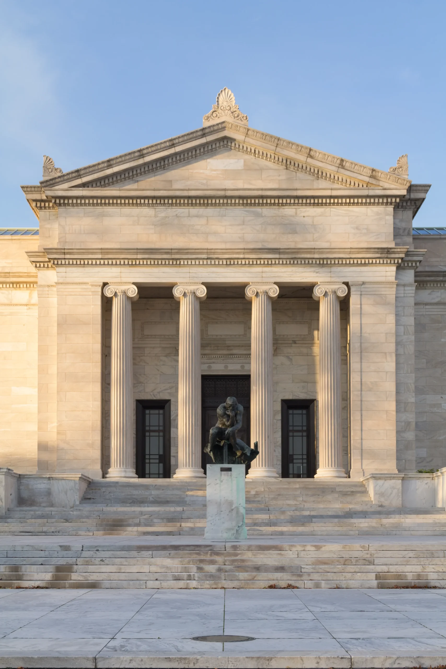 A bronze statue of a pensive, nude man sits on a pedestal at the base of a wide staircase leading to a grand classical stone building with four columns.