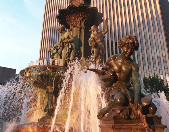 A bronze multi-tiered fountain sprays water vigorously, featuring statues like a foreground male figure and a top female figure with outstretched arms, against a striped skyscraper.
