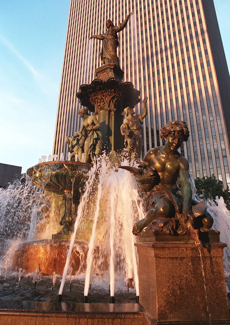 A bronze multi-tiered fountain sprays water vigorously, featuring statues like a foreground male figure and a top female figure with outstretched arms, against a striped skyscraper.