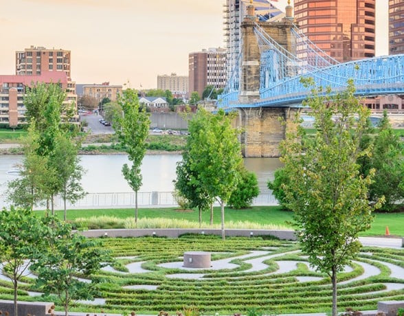 A landscaped urban park features a circular green maze pattern with a central stone, overlooking a river, a blue suspension bridge, and a city skyline.