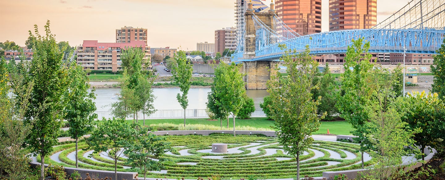 A landscaped urban park features a circular green maze pattern with a central stone, overlooking a river, a blue suspension bridge, and a city skyline.