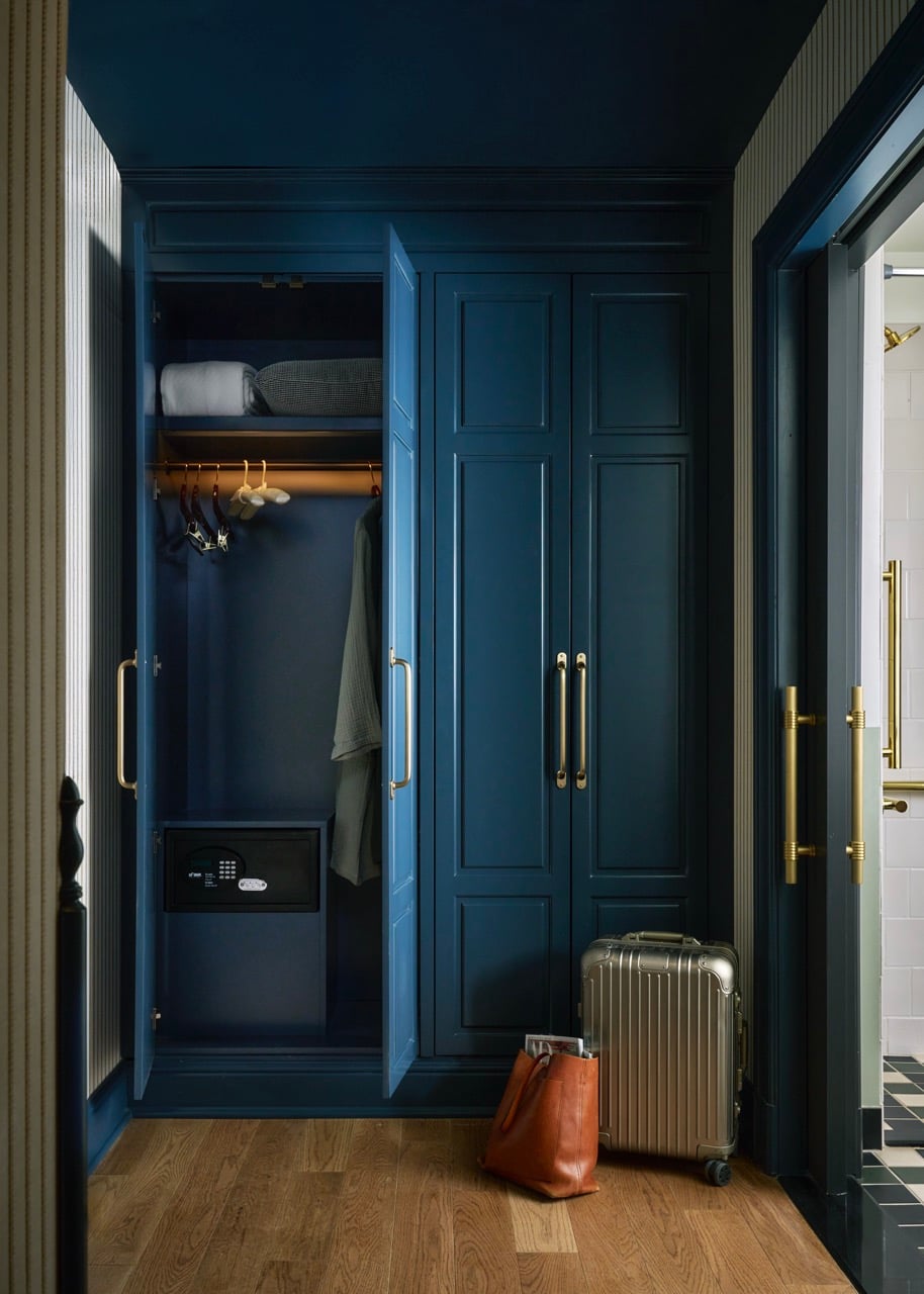 Dark blue wardrobe with its left door ajar, revealing a safe labeled "SäFE XTRAM", hangers, and folded items. A silver suitcase and brown bag sit on the wooden floor beside it, near a bathroom doorway.