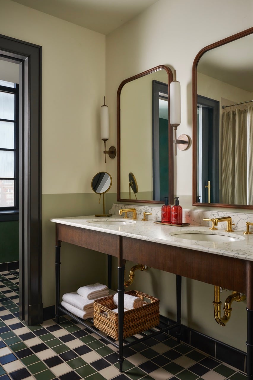Two-sink vanity, gold faucets, mirrors, and two red "FIDELITY" bottles. The bathroom features two-tone walls and a green/black/cream checkerboard floor.