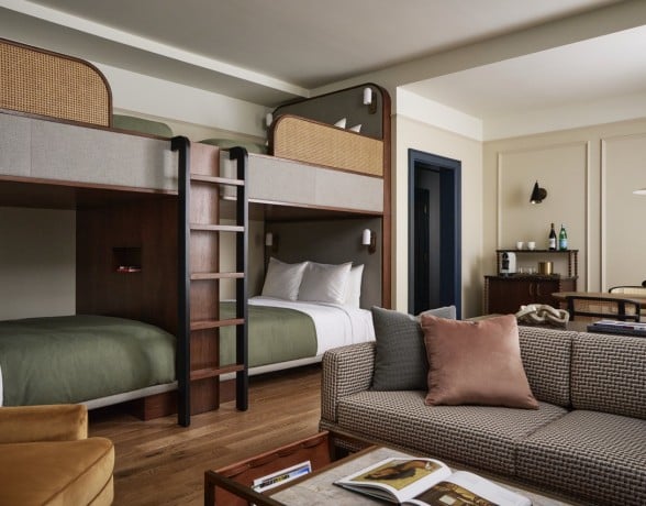 Modern dark wood bunk beds with green bedding and cane accents feature in a stylish room, alongside a patterned sofa, coffee table, and mini-bar, all on hardwood floors.