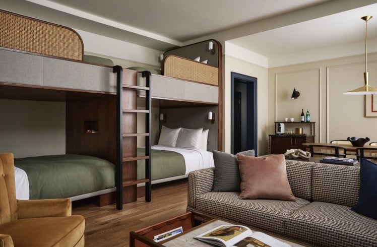 Modern dark wood bunk beds with green bedding and cane accents feature in a stylish room, alongside a patterned sofa, coffee table, and mini-bar, all on hardwood floors.
