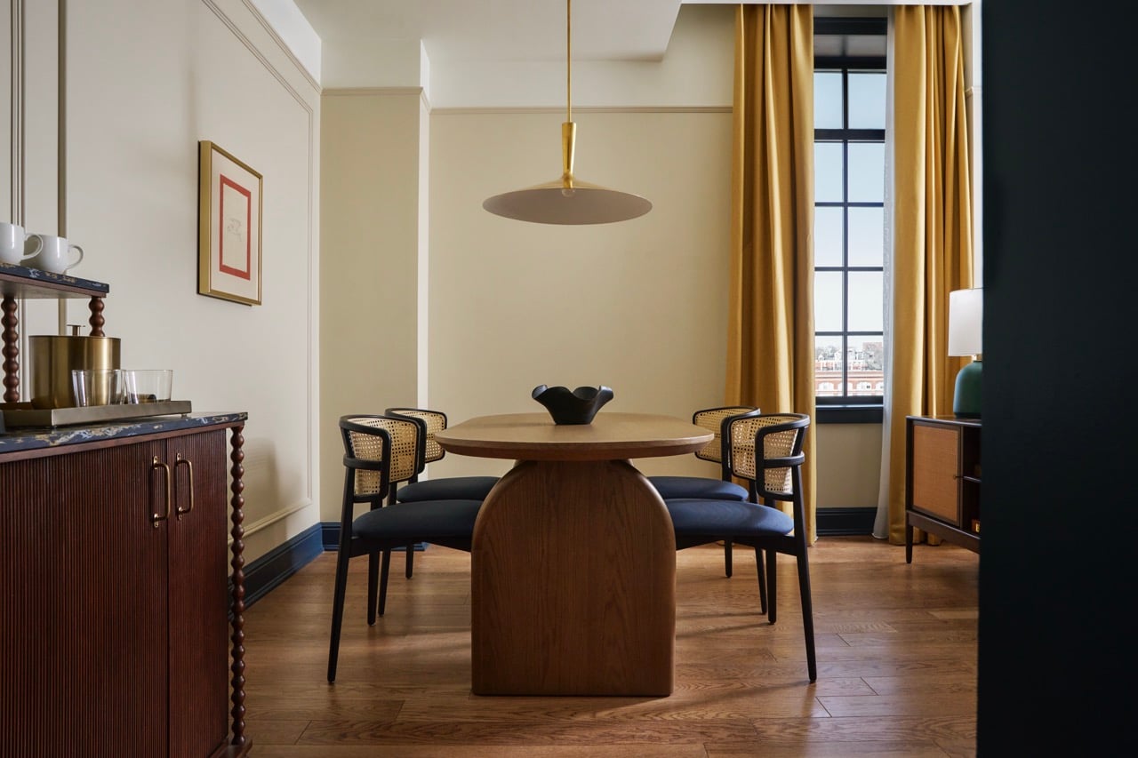A modern wooden dining table surrounded by four cane-backed chairs sits on hardwood floors. A gold lamp hangs above; a bar cart and window with gold curtains complete the room.