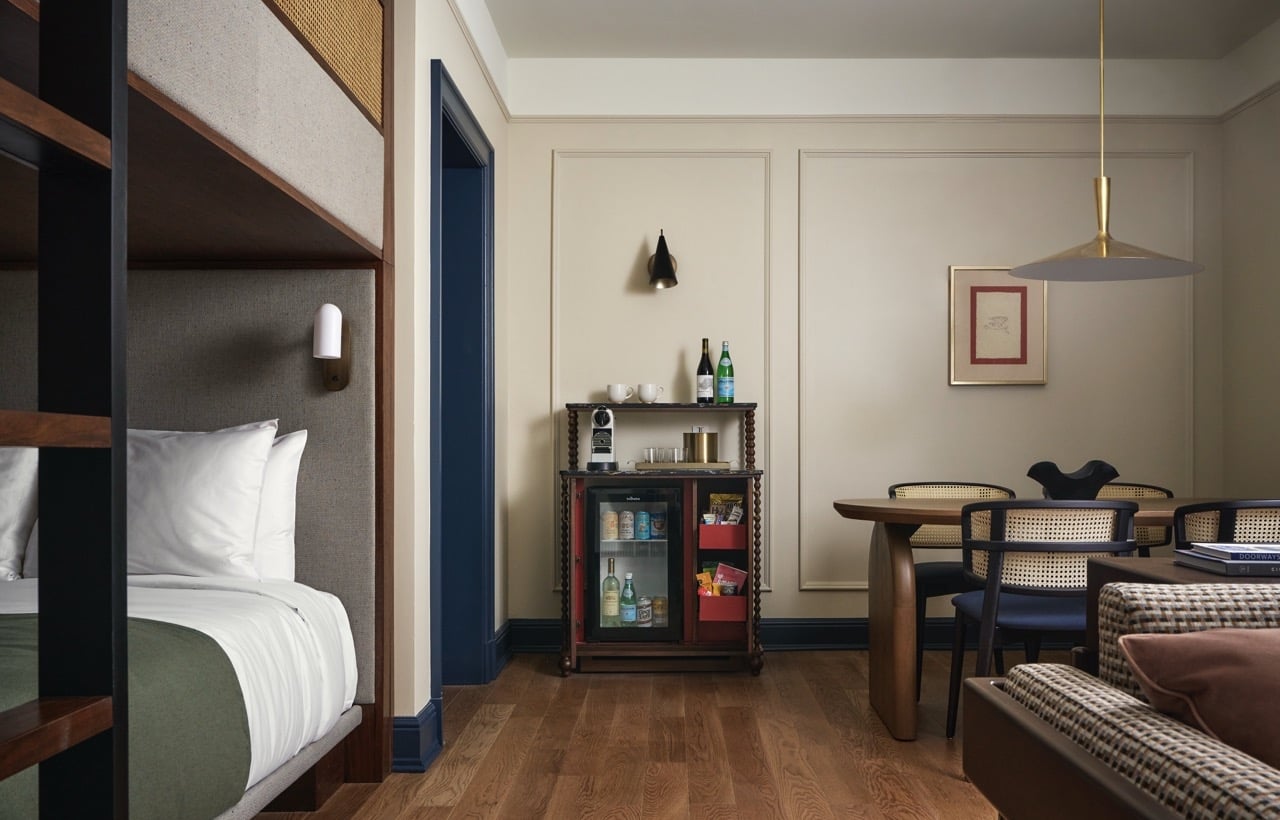 A room features a lower bunk, a minibar with fridge/coffee maker, and a dining table. Light walls, wooden floors define the space, with "DOORWAYS" on a couch.