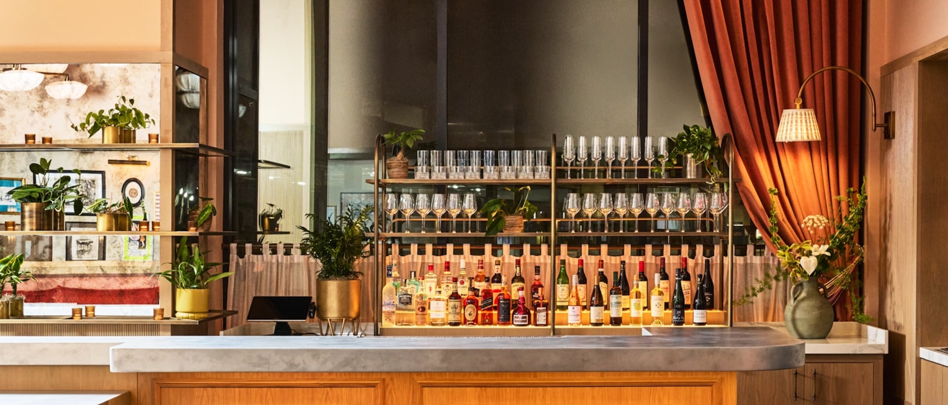 A fully stocked bar displays liquor and glassware within a warmly lit restaurant. Set dining tables, dark green bar stools, and ornate chandeliers complete the inviting ambiance.