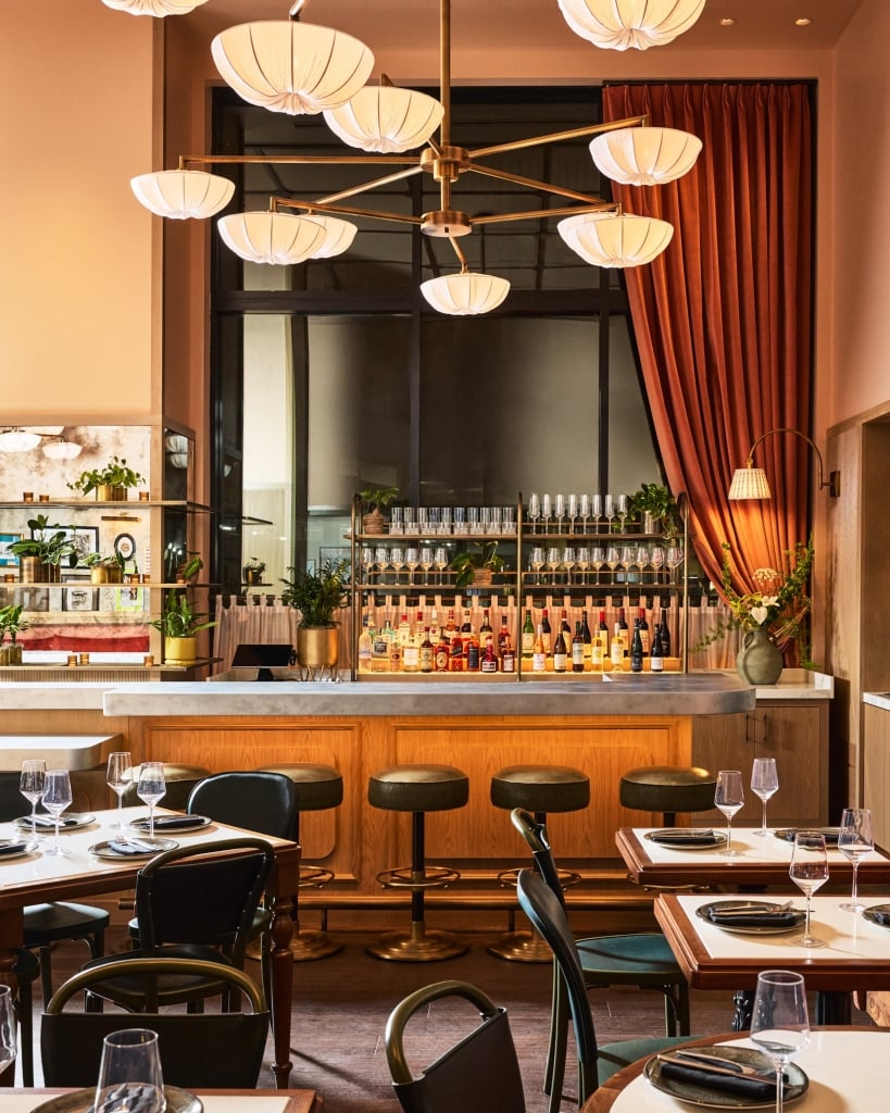 A fully stocked bar displays liquor and glassware within a warmly lit restaurant. Set dining tables, dark green bar stools, and ornate chandeliers complete the inviting ambiance.