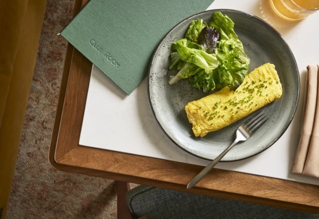 An omelette with chives and a side salad are served on a gray plate with a fork, on a marble-topped wooden table. A green menu reading "CLUB ROOM" and a drink are also present.