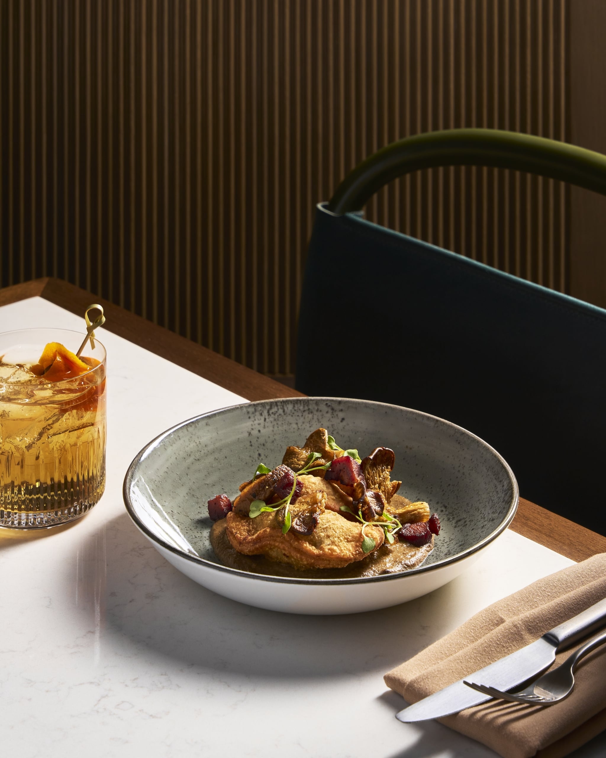 A dish of golden-fried dumplings with mushrooms, red cubes, and microgreens in a grey bowl, next to a garnished cocktail, on a white marble table.