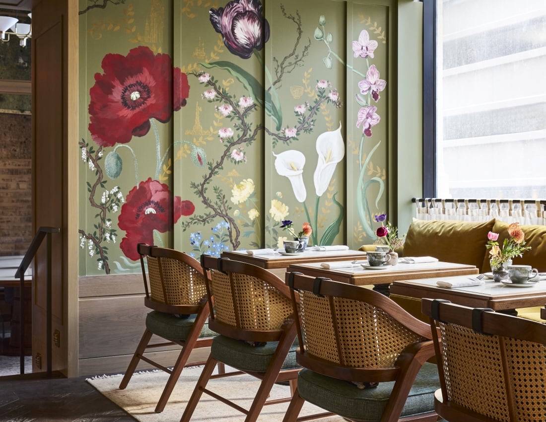 Chic dining area with dark wood, cane-back chairs and set tables. Olive-green walls feature vibrant floral murals (poppies, orchids) and a matching patterned ceiling, by a sunlit window.