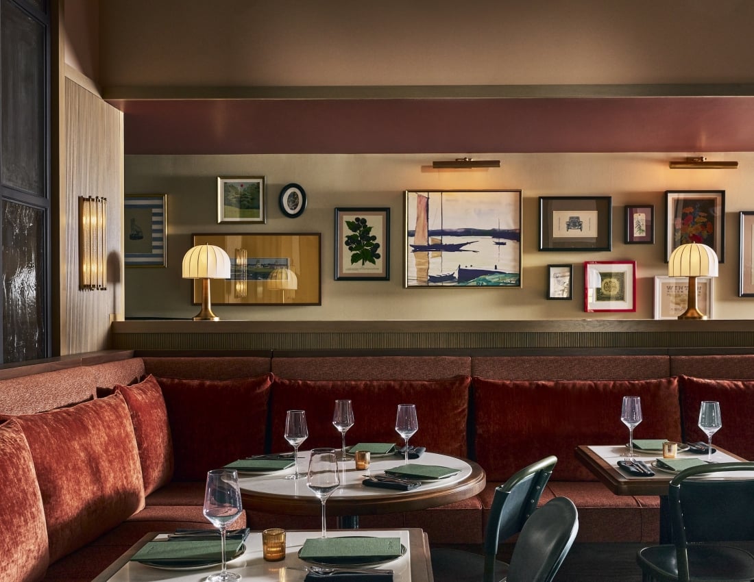 Restaurant interior with a long red velvet banquette and tables set for dining. Walls display various framed artworks under warm lighting.