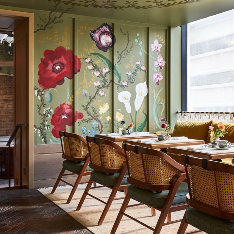 Dining room features several set tables and wooden chairs with cane backs. Green walls display vibrant floral murals, while the ceiling has an intricate leaf pattern.