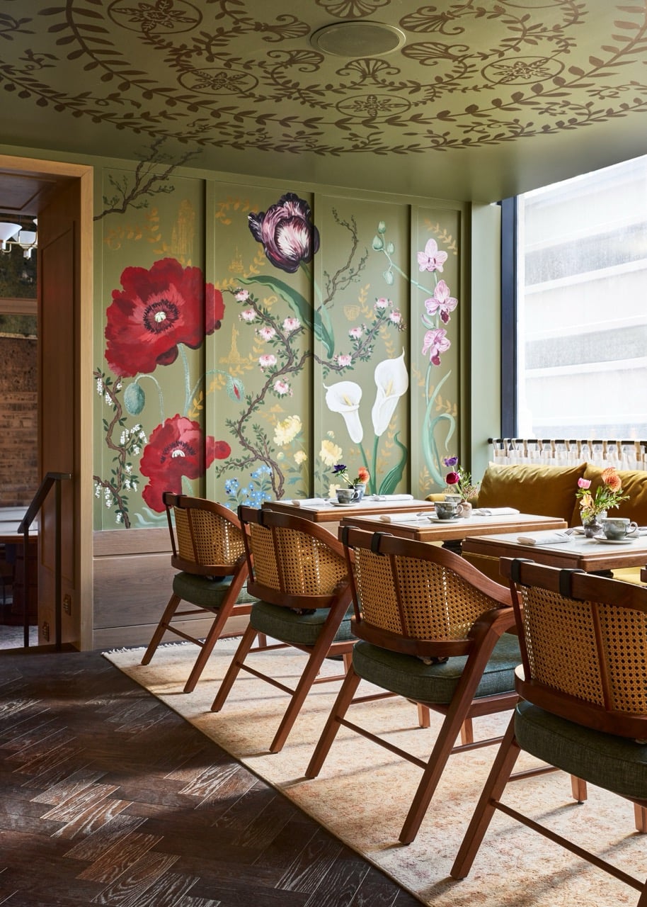 Dining room features several set tables and wooden chairs with cane backs. Green walls display vibrant floral murals, while the ceiling has an intricate leaf pattern.