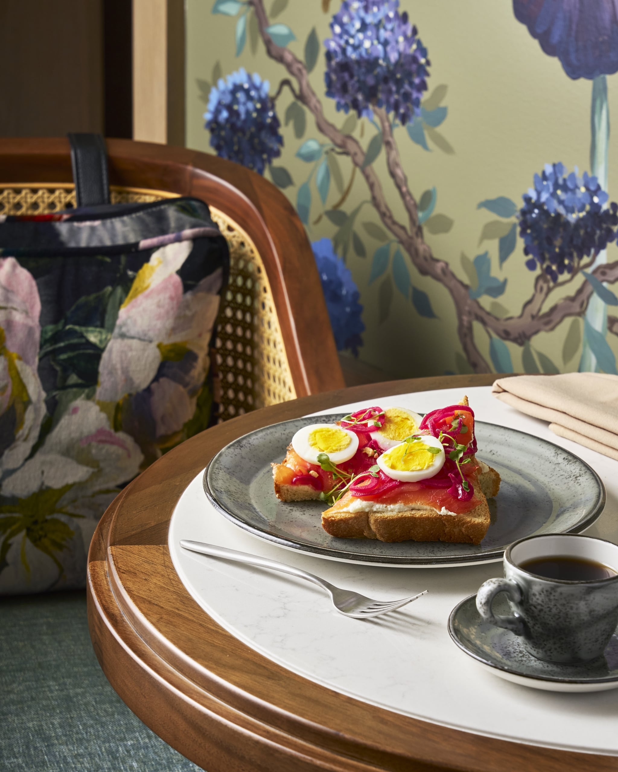 Two open-faced toasts with smoked salmon, pickled onions, microgreens, and halved eggs, with a coffee cup and fork, on a table against a chair and floral wallpaper.
