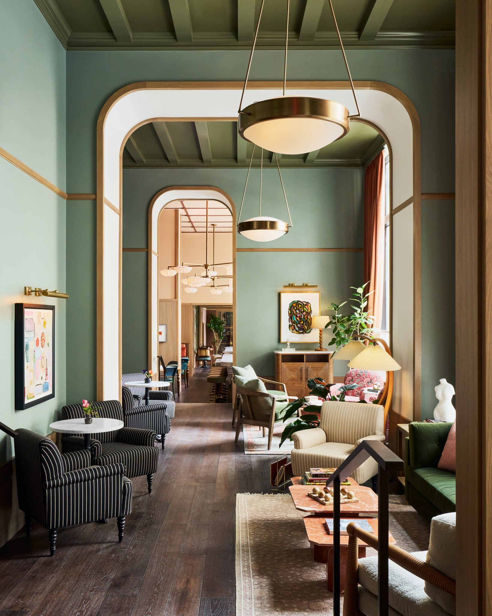 A long, elegant hotel lounge features sage green walls, dark wood floors, arched doorways, and diverse seating arrangements with brass lighting, extending into further rooms. Text: "Frida Kahlo CLASSIC AMERICA".