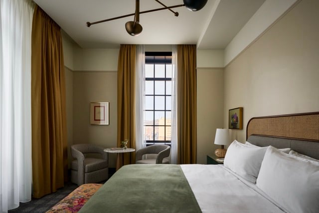 A neatly made bed with white pillows and a green blanket fills the foreground of a hotel room featuring two armchairs, a small table, and a tall window with brown curtains.
