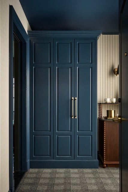 Dark blue built-in wardrobe with brass handles in an entryway. It features striped walls, a dark blue ceiling, and plaid carpet. A wooden table with cups is to the right.