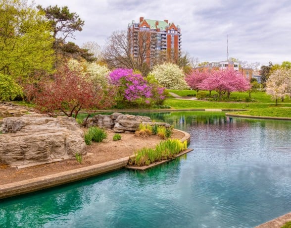 Eden Park in spring with a curved turquoise pond, large rocks, and blooming pink, white, and purple trees under a cloudy sky. A tall residential building rises in the background beyond the green lawns and walking paths.